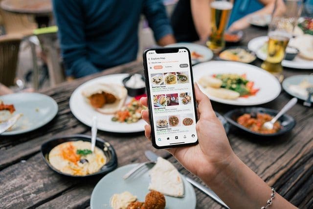 Pessoa olhando cardápio de restaurante no celular. Atrás de sua mão, há pessoas se servindo com comidas na mesa.