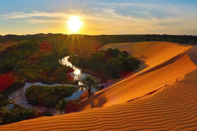 Vista do topo das dunas do Jalapão, no Tocantins, localizadas à direita da foto, e vista da vegetação nativa à esquerda e também ao fundo. O sol está se pondo e está acima da vegetação, refletindo nas dunas e dando um tom alaranjado.