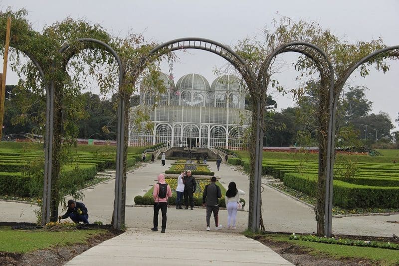 Foto de um portal que dá acesso ao jardim e à estufa do Jardim Botânico, em Curitiba, no Paraná. Embaixo do portal, estão algumas pessoas, que passam por ele para acessar o jardim.