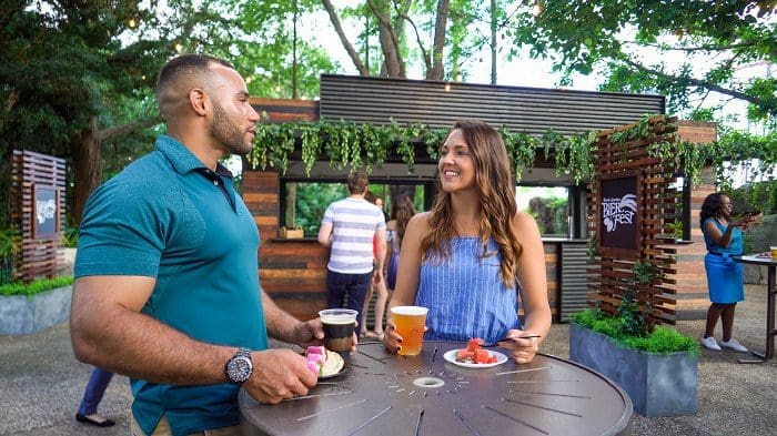 Pessoas conversando, segurando copos de bebidas em cima de uma mesa marrom, ao lado de um prato, no Bier Fest, no Busch Gardens Tampa Bay, na Flórida. Ao fundo, há uma lanchonete de madeira, com plantas verdes em cima.