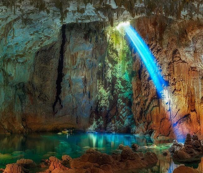 bonito_em_ms_foto_marcio_cabral_photography_site_acqua_studio_de_viagens Vista de uma caverna de Bonito, no Mato Grosso do Sul. A caverna possui formações rochosas em tom marrom alaranjado, água em tons azul turquesa e verde, uma abertura superior, pela qual passa a luz do sol e reflete a luz azul turquesa na pedra.