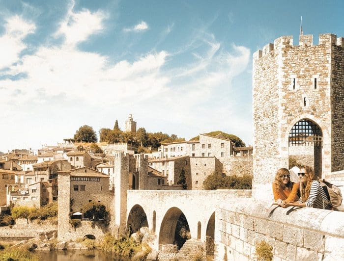 cidade_de_catalunha Cidade de Catalunha, na Espanha, mostrando a arquitetura europeia espanhola, que se assemelha à castelos e também à arquitetura romana. Á direita da foto, duas mulheres estão com os braços apoiados no parapeito de uma construção, admirando a vista. O céu é azul claro, com algumas nuvens.