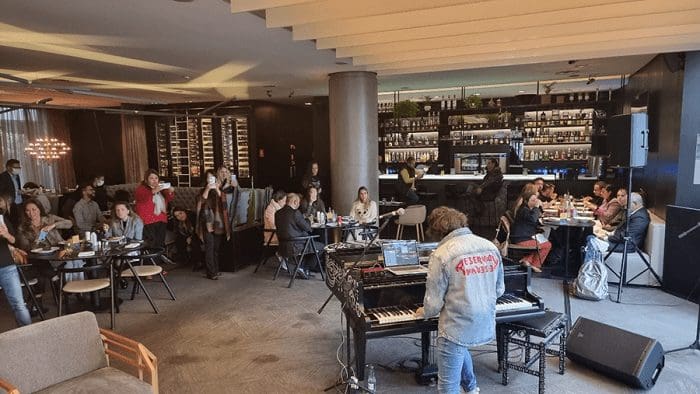 Evento Rock´n Taste IT, no Pullman Ibirapuera. Há algumas pessoas comendo na mesa, um músico tocando piano, e outas pessoas, à esquerda, assistindo.