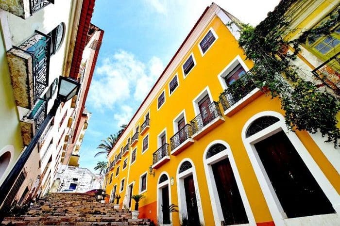Casarões em São Luís - MA. O casarão em destaque está no lado direito, é no estilo colonial, em amarelo. A rua é de paralelepípedos. Do lado esquerdo e no fundo da foto, as casas são brancas.