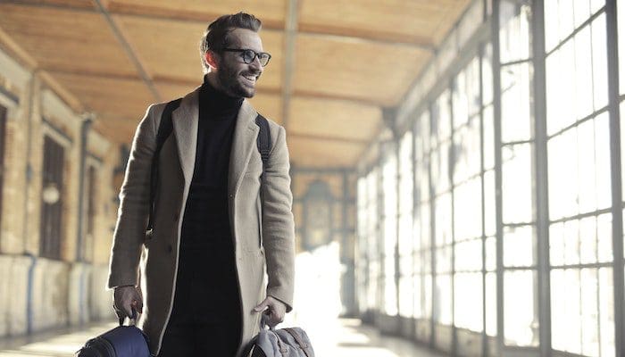 pexels-andrea-piacquadio-837129 Homem loiro de óculos, usando sobretudo e segurando duas malas nas mãos e uma mochila nas costas, está sorrindo e fará uma de suas viagens a trabalho.