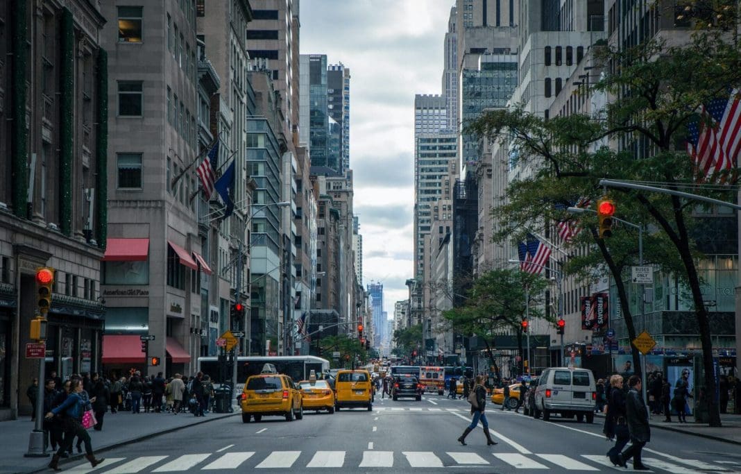 pexels-nout-gons-378570 Uma das ruas da cidade de Nova York, nos Estados Unidos. Há táxis amarelos no trânsito, prédios com bandeiras dos Estados Unidos hasteadas, e uma pessoa atravessando a rua.