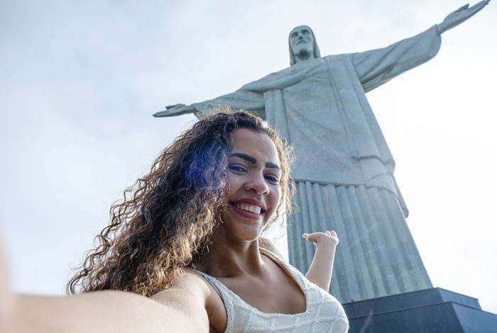 Mulher sorridente faz uma selfie e mostra o Cristo Redentor, no Rio de Janeiro, ao fundo. A 123milhas fez uma parceria com a ETS e oferece serviços turísticos em sua plataforma.