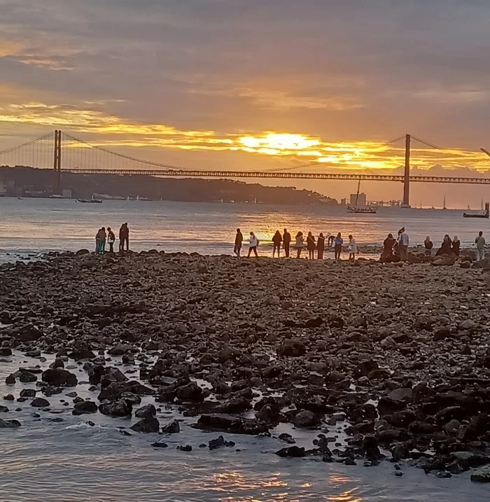 Imagem de uma praia de pedras, em Lisboa, capital de Portugal, com algumas pessoas.