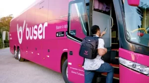 Passageiro negro, de costas, embarcando no ônibus da Buser, com uma mochila da Buser nas costas. A cor do ônibus é rosa. 10 viagens de ônibus mais reservadas em 2022: Buser divulga lista.