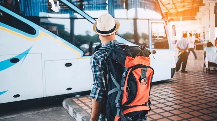Viajante de costas usando camisa xadrez azul, com mochila nas costas laranja e usando um chapéu branco com fita preta. Ele olha para um ônibus. 