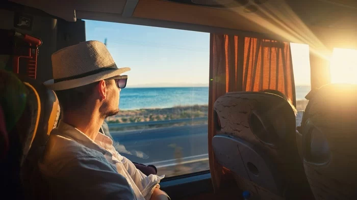 Viajante de perfil, sentado dentro de um ônibus, olhando a janela. Por ela, ele enxerga a estrada e ao fundo, o mar azul. Ele usa óculos escuros, camisa branca clara e um chapéu branco com fita preta. A 123ônibus foi lançada pela 123milhas.
