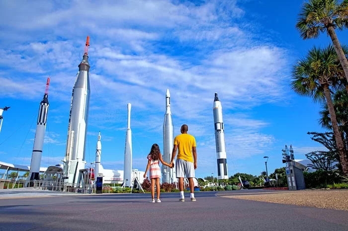Pai e filha negros, de costas e em roupas casuais, na entrada do Kennedy Space Center Vistor Complex, de dia, admirando foguetes.