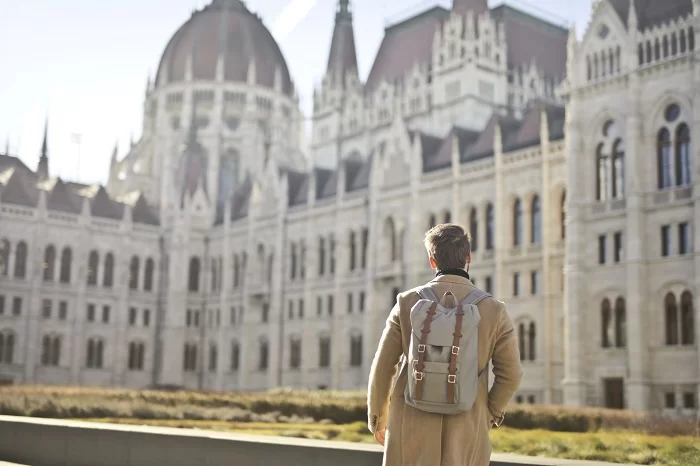 male-wearing-brown-coat-backpack-near-hungarian-parliament-building-budapest-hungary Homem de costas, com roupa social e mochila, vendo um castelo na Alemanha, de dia. Turismo na Alemanha: veja quanto se gasta com saúde em casos de emergência e a importância do seguro viagem